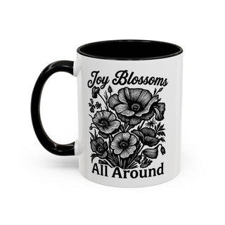 White mug with black handle, featuring "Joy Blossoms All Around" text and poppy illustration.